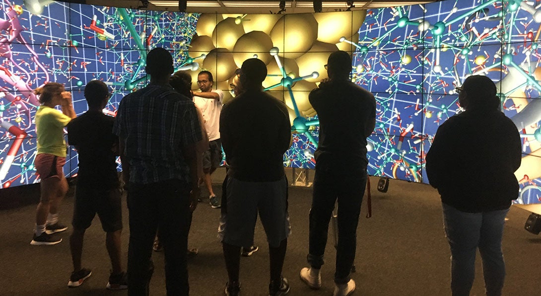 A small group of students stand listening to a presenter who stands in front of a wall of screens with colorful abstract graphics that look somewhat like molecules.